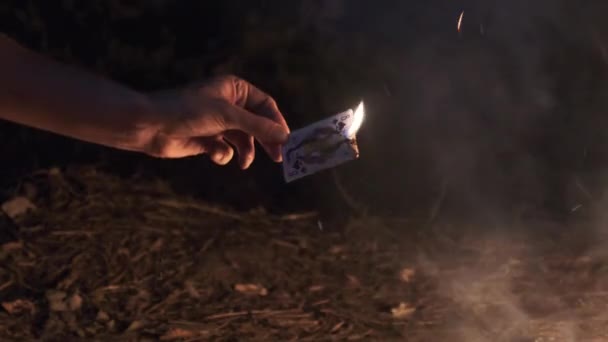 Hand håller brinnande kort på svart bakgrund nära Campfire on Nature. Långsamma rörelser — Stockvideo