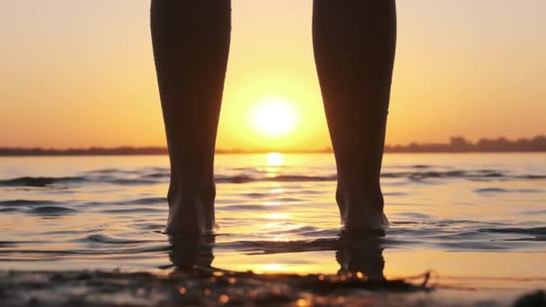 Silhueta de pernas de mulher em pé na água ao pôr do sol na praia. Movimento lento — Vídeo de Stock