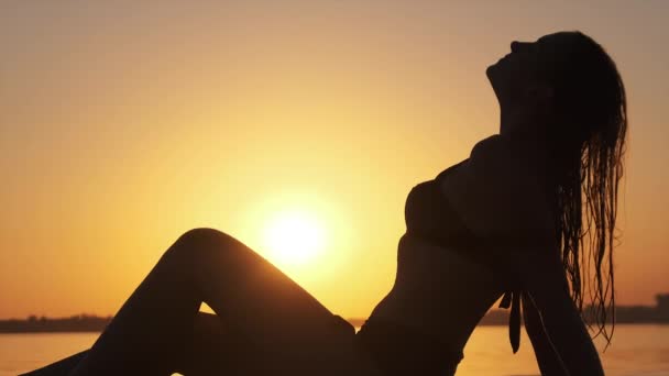 Silhouette einer heißen Frau im Badeanzug, die am Abendstrand bei Sonnenuntergang liegt und posiert — Stockvideo