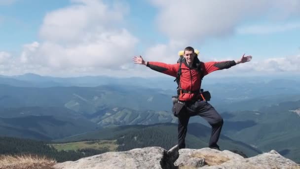 Turista com mochila no topo da montanha perto do penhasco levanta as mãos para o lado — Vídeo de Stock