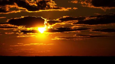 Altın güneş, karanlık, siluetli bulutların arkasından inerken, ufuktaki turuncu gökyüzünün üzerinde güzel güneş ışınları yayarken, zaman kavramı muhteşem bir gün batımı yakalar..
