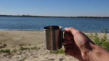 Bir gezgin parlak bir günde kumlu göl kıyısının manzaralı arka planında metal bir kamp kupası tutuyor. Bir açık hava macerası sırasında ya da deniz kenarında bir yolculuk sırasında dinlenme anı..