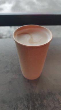 Masadaki kağıt bardakta latte tabağıyla servis edilen kahvenin dikey görüntüsü. Arka planda, insanlar geçerken bulanık bir şehir caddesinin pencerenin içinden geçerek rahat bir kafe havası yarattığını görüyoruz..