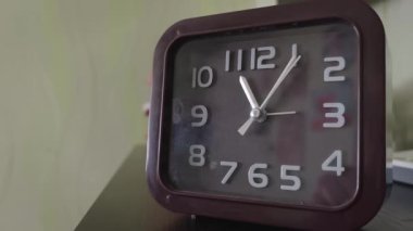 Kare kahverengi analog alarm saati, üzerinde beyaz numaralar olan siyah bir masada durur. Kamera yavaşça uzaklaşıyor. İkinci el sürekli hareket ediyor. Zamanın akışını gösteriyor..