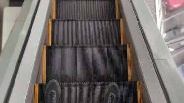 Yürüyen merdivenden aşağı inerken siyah ayakkabılarla ayaklara bakan bir bakış açısı. Hareket halindeki merdivenlerden en alt kattaki metal iniş kaplamasına adım atan kişi..