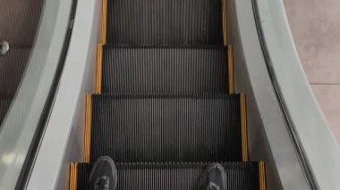 Siyah ayakkabılarla yürüyen merdivene binen ayakları gösteren bir pov fotoğrafı. Bir sonraki kata varır ve merdivenlerden inip modern bir binanın metal iniş kaplamasına çıkar..