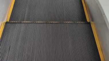 Yakından, yüksek açılı boş yürüyen merdiven manzaralı. Sarı güvenlik çizgileri olan yivli metal basamaklar alçaldıkça hipnotik, mekanik desenler yaratır ve kaybolurlar..