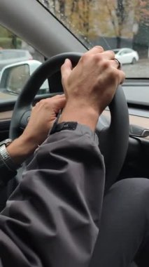 Kasvetli bir günde bir adamın direksiyondaki ellerini şehrin sokaklarında gezdirirken görüntüsü. Dikey perspektif sürüş eylemine ve şehir içi ulaşıma odaklanıyor.