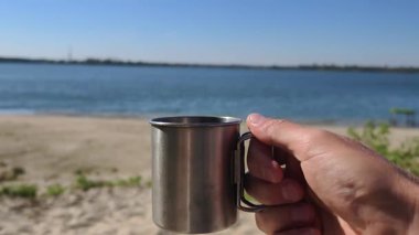 Kumlu bir sahilde paslanmaz çelik bir seyahat kupasını tutan bir elin birinci şahıs görüntüsü. Arka planda, sakin mavi bir göl ve açık bir gökyüzü sakin bir açık hava kampı ya da piknik sahnesi oluşturur..