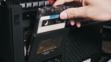 Klasik bir teyp destesini açan bir elin yan açısı görüntüsü, mavi etiketli klasik bir ses kaseti takmak, kapıyı kapatmak ve müziği başlatmak için gümüş çalma düğmesine basmak.