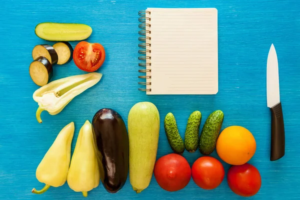 Vegetables are whole and cut a notepad for writing ricepts and a ceramic knife on a blue board.