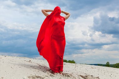 Kumda kırmızı bir bez giymiş bir kız bulutlu bir gökyüzünün arkaplanına karşı bir fotoğrafçıya poz veriyor. Rüzgardaki kırmızı kumaş kızın vücuduna sarılıyor..