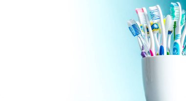 Multi-colored toothbrushes in a white mug close-up with a gradient background from white to blue with an isolated place for text