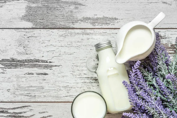 Vintage glass bottle with milk, a glass with milk and a beautiful white jug next to a branch of lavender flowers on a vintage wooden background. View from above.