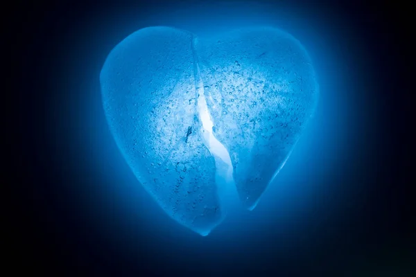 Ghiaccio congelato rotto a metà cuore simbolo di colore blu primo piano incandescente al buio in luce fredda blu. Texture di ghiaccio con bolle. Simbolo di un cuore spezzato. Allegoria dell'amore infelice . — Foto Stock