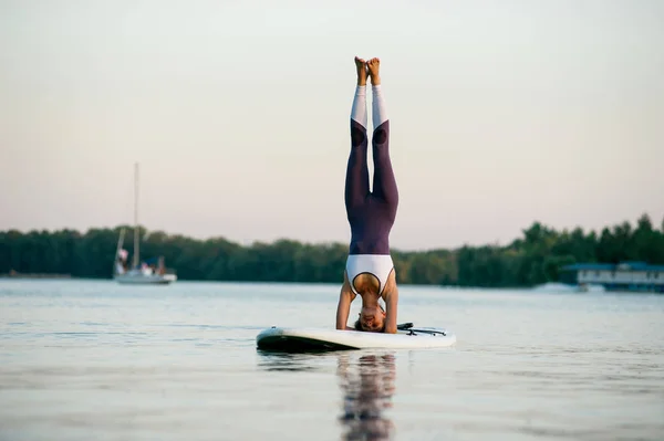 stand up paddle board yoga performed by beautiful girl on the bright city background, girl is standing on her head
