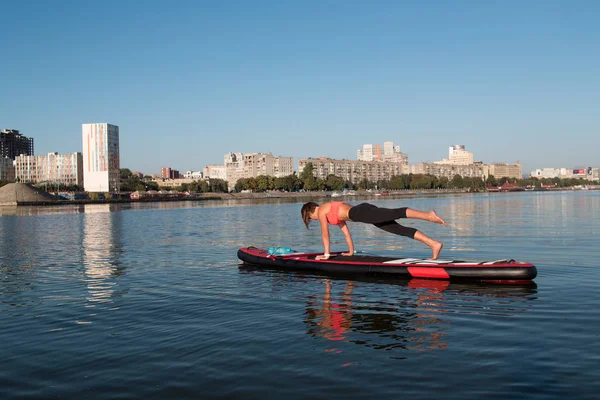 stand up paddle board yoga performed by beautiful girl on the bright city background, yoga training on the water