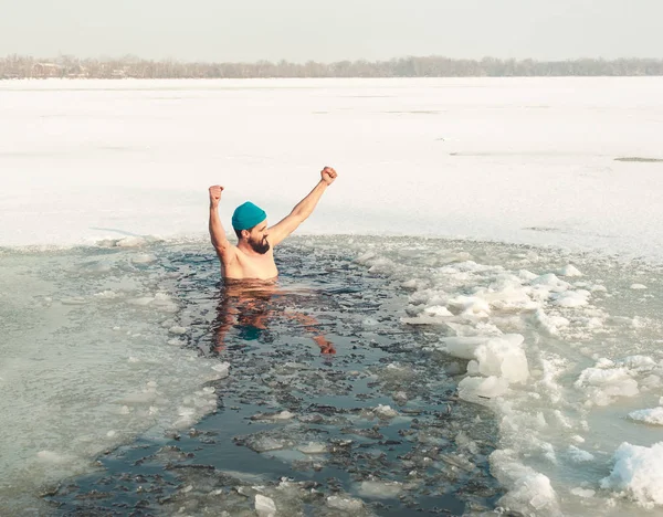 bearded man in hat swimming in the winter lake in the ice hole, ice hole swimming, raised his hands up and looks away