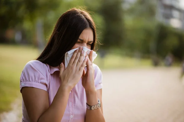 Woman with allergy symptom blowing nose in park.