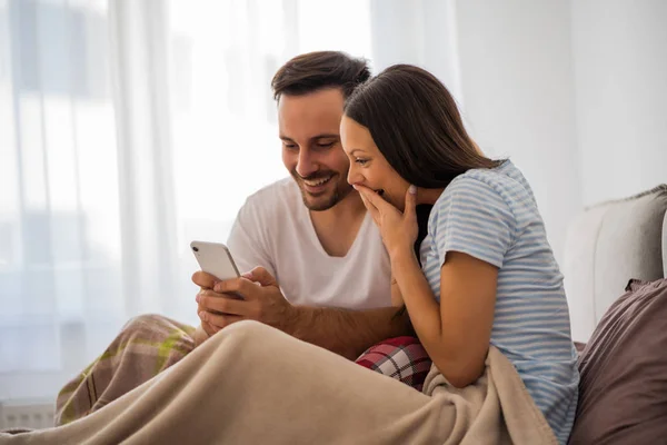 若い幸せなカップルは 朝ベッドでスマートフォンを見ています — ストック写真