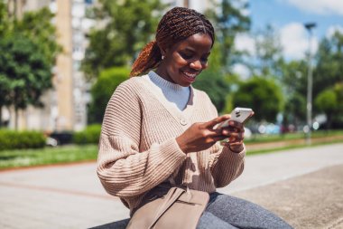 Telefonuyla mesajlaşırken gülümseyen Afro-Amerikan kadın, gün ışığında güneşleniyor, boş zamanları ve iletişimi sembolize ediyor.