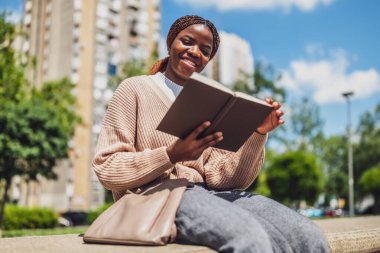 Neşeli Afrikalı Amerikalı kadın şehir parkında dinleniyor, kitap okuyor..