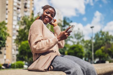 Şehir ortamında cep telefonunu kullanan, kablosuz kulaklık takan, arkadaşlarına mesaj atarken gülümseyen, modern şehir hayatını ve boş zamanlarını somutlaştıran, 20 'li yaşlarındaki neşeli siyahi bir kadının portresi..