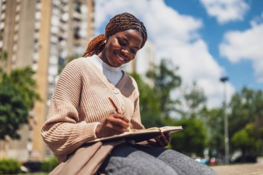 Neşeli Afrikalı Amerikalı kadın şehir parkında dinleniyor, günlüğünü yazıyor..