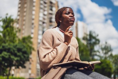 Afro-Amerikalı kadın şehir parkında dinleniyor, derin düşüncelere dalmış ve günlüğünü yazıyor..
