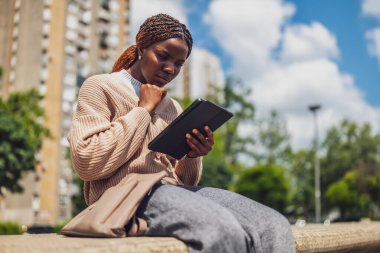 20 'li yaşlarda Afro-Amerikalı bir kadın tabletiyle etkileşime girerken endişeli ve hoşnutsuz görünüyor. Açık havada oturuyor ve şehir hayatının baskılarını yansıtıyor..