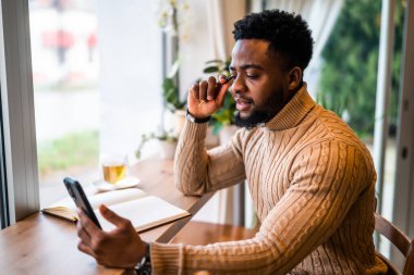 Barda oturan genç siyahi adam. Akıllı telefon ve not defteri kullanıyormuş. Serbest İşadamı iş planları yapıyor.