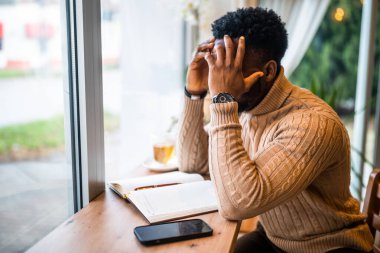 Barda oturan genç siyahi adam. Akıllı telefon ve not defteri kullanıyormuş. Serbest İşadamı iş planları yapıyor.