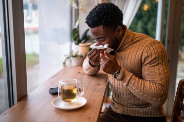 Genç adam barda oturuyor, rahatlıyor ve çay içiyor. Burnunu siliyor..