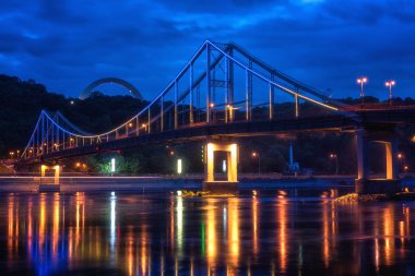 Ukrayna 'nın başkenti Kyiv (Kiev), renkli ışıklarla ve sudaki yansımasıyla Avrupa' nın gece kenti. Dinyeper nehri boyunca yaya köprüsü ve nehir istasyonu manzarası.