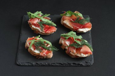 Keçi peynirli karabuğday tostları, prosciutto ve roka siyah bir masada siyah bir taş tahtada yatıyor. Yakın çekim.