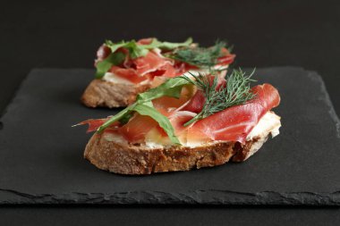 Keçi peynirli karabuğday tostları, prosciutto ve roka siyah bir masada siyah bir taş tahtada yatıyor. Yakın çekim.
