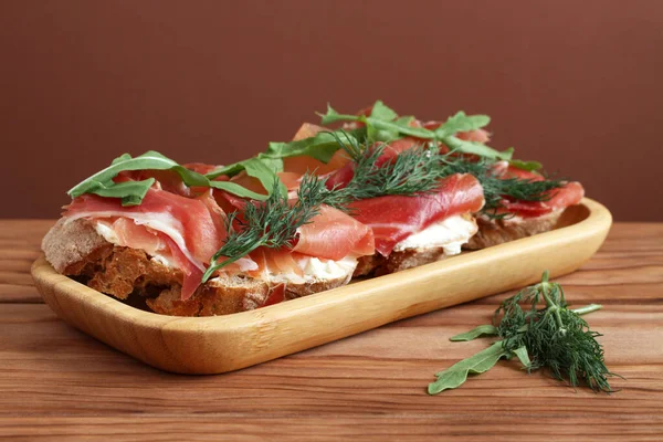 Tahta bir masada bambu tabağında duran keçi peyniri, jambon ve roka ile karabuğday tostları. Yakın çekim.