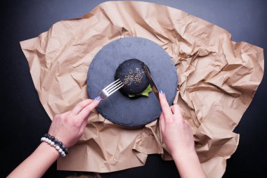 Siyah susamlı siyah ekmek rulosu insan eli siyah arka planda bir kraft kağıdının üzerinde duruyor. Garip siyah hamburger ekmeğinin üstünde. Cadılar Bayramı yemeği