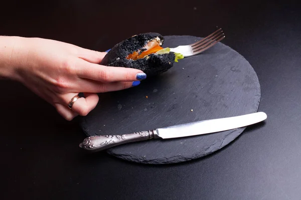 Rollo de pan negro con sésamo negro sostenido en las manos sobre un papel kraft en manos humanas fondo negro. Por encima de la vista de pan de hamburguesa negro extraño. Comida Halloween —  Fotos de Stock