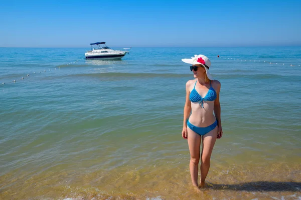 一个戴着墨镜和白帽子的金发女郎站在游艇对面的海滩上 — 图库照片