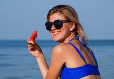 Güneş gözlüklü sarışın deniz kenarında bir karpuz yiyor. Bir kadın elinde sulu bir karpuz