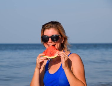 Güneş gözlüklü sarışın deniz kenarında bir karpuz yiyor. Bir kadın elinde sulu bir karpuz
