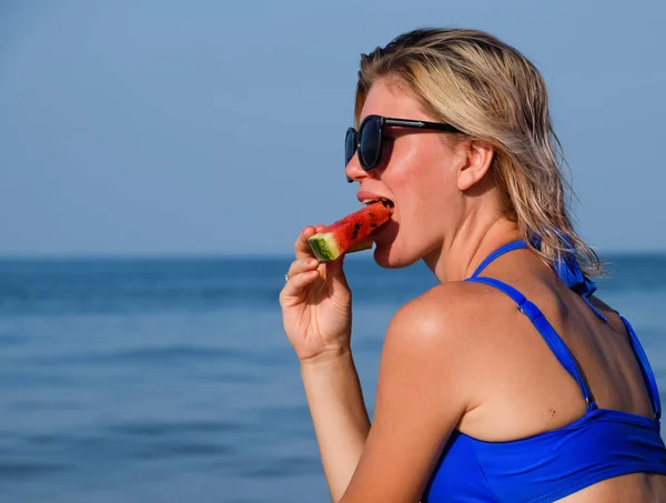Uma loira de óculos de sol come uma melancia junto ao mar. Uma melancia suculenta nas mãos de uma mulher — Fotografia de Stock