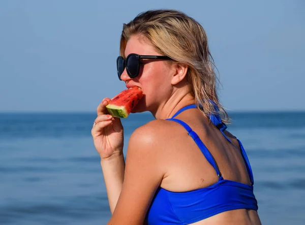 Uma loira de óculos de sol come uma melancia junto ao mar. Uma melancia suculenta nas mãos de uma mulher — Fotografia de Stock