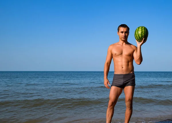 Ένα σπορ άνθρωπος στέκεται στην παραλία και κρατώντας ένα ολόκληρο καρπούζι. Ένα ώριμο καρπούζι στα χέρια ενός ανθρώπου — Φωτογραφία Αρχείου