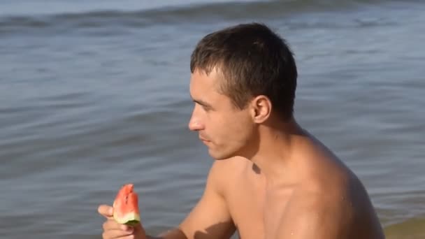 Un hombre está comiendo una sandía sentada en la playa. Una sandía madura en las manos de un hombre — Vídeos de Stock
