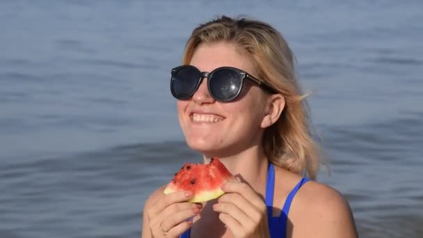 一个戴墨镜的女人吃了坐在沙滩上的西瓜。一个成熟的西瓜在一个金发女郎的手中 — 图库视频影像