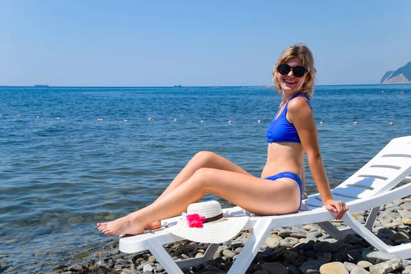 Menina bonita em um biquíni azul e chapéu branco banhos de sol na praia, sentado em um salão de chaise branco. Loira com óculos de sol — Fotografia de Stock