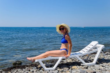 Güzel kız mavi bikini ve beyaz şapka beyaz şezlong üzerinde oturan sahilde güneşlenmek. Sarışın gözlüklü