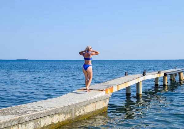 Красивая девушка в синем бикини идёт к морю. Бетонный причал. Прыжки в воду с пирса. Девчонки — стоковое фото